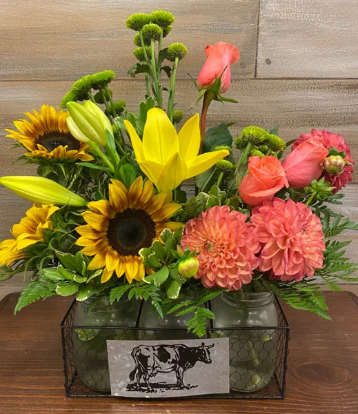 A floral arrangement with sunflowers, yellow lilies, and pink roses in a wire basket with a cow silhouette tag.