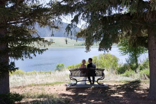 A bride and groom sitting on a bench overlooking a lake, framed by evergreen trees.