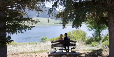 A bride and groom sitting on a bench overlooking a lake, framed by evergreen trees.