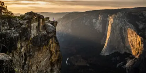 A couple standing on a rocky ledge overlooking Yosemite Valley at sunset.