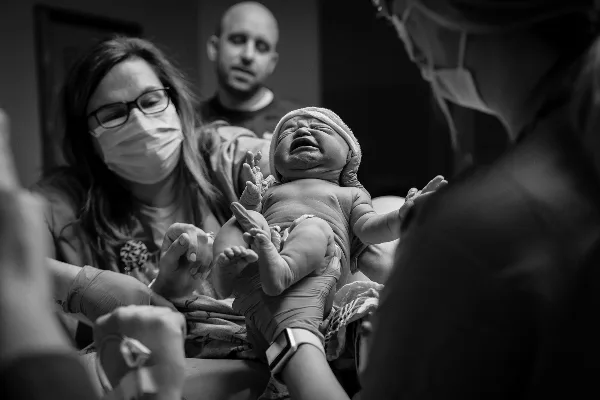 A black-and-white photograph of a crying newborn baby being held by a mother in a hospital setting, with a father in the background.