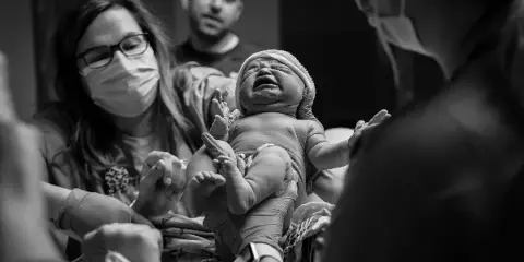 A black-and-white photograph of a crying newborn baby being held by a mother in a hospital setting, with a father in the background.