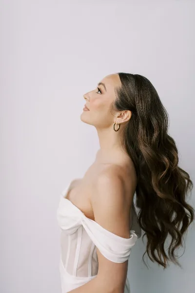 Bride in profile with styled wavy hair and makeup, wearing a white off-shoulder dress.