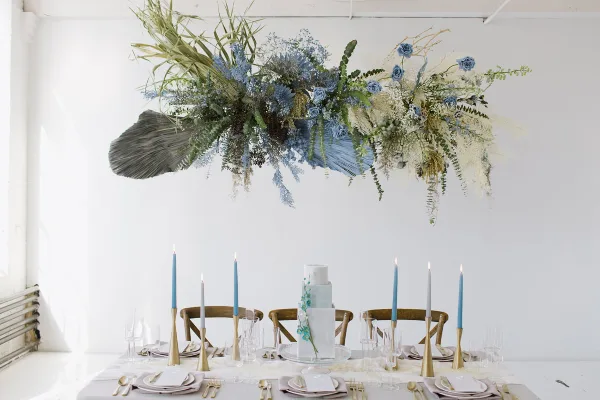 A white and light blue tiered wedding cake with floral accents displayed on a table with blue candles and a large floral installation.