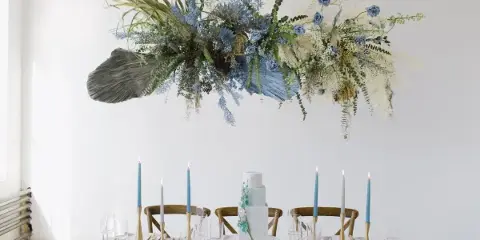 A white and light blue tiered wedding cake with floral accents displayed on a table with blue candles and a large floral installation.