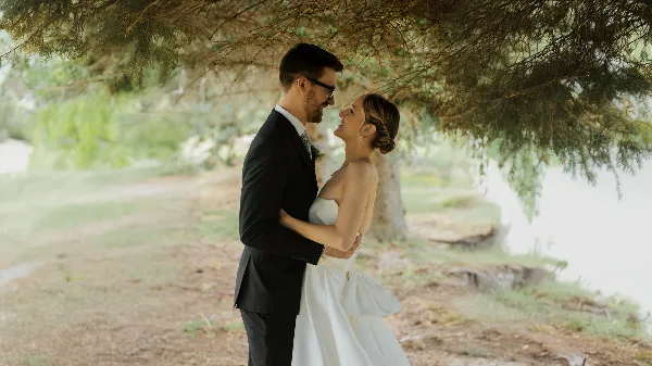 A bride and groom embracing and smiling at each other outdoors under a large tree.