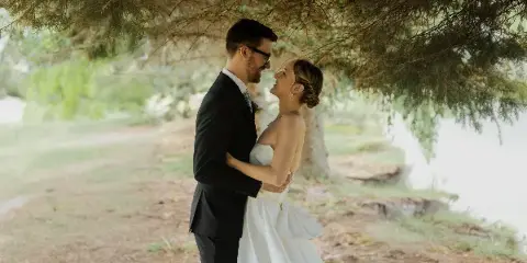 A bride and groom embracing and smiling at each other outdoors under a large tree.