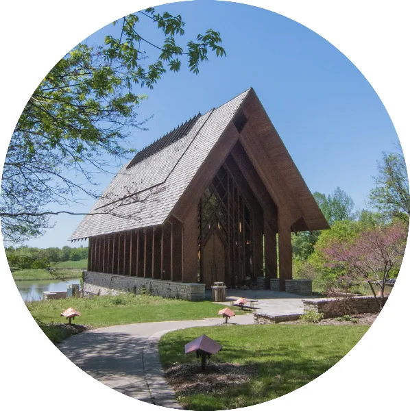 Exterior view of the Marjorie Powell Allen Chapel, a wooden A-frame structure with a steep shingled roof, situated on a grassy lawn near a body of water.