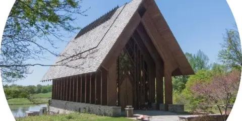 Exterior view of the Marjorie Powell Allen Chapel, a wooden A-frame structure with a steep shingled roof, situated on a grassy lawn near a body of water.
