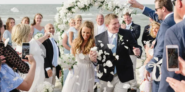 A bride and groom walking down the aisle at a beach wedding ceremony under a white floral arch with guests throwing petals.