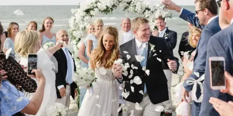 A bride and groom walking down the aisle at a beach wedding ceremony under a white floral arch with guests throwing petals.