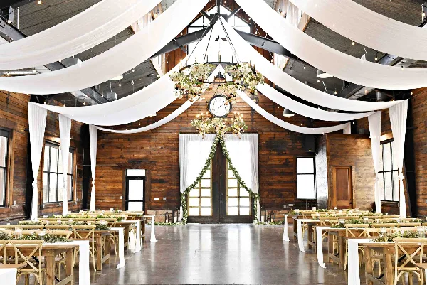 Interior of a rustic wooden barn wedding venue with white ceiling drapery, floral chandelier, and reception tables.