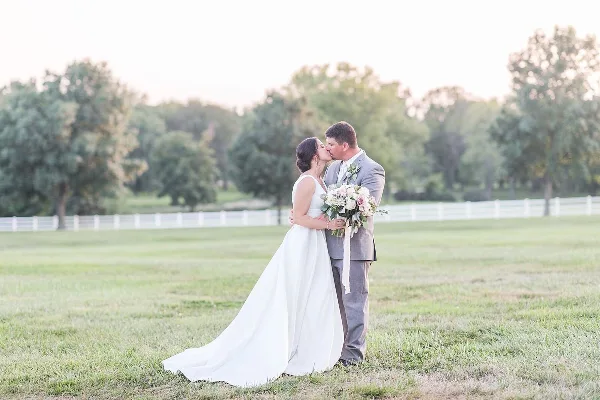 A bride and groom kissing in a grassy field with a white fence and trees in the background.