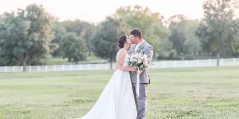 A bride and groom kissing in a grassy field with a white fence and trees in the background.