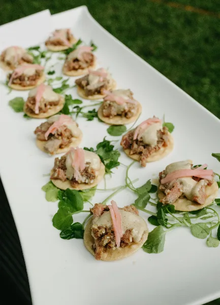 A white rectangular platter holding a row of gourmet appetizers topped with meat, cheese, and garnished with fresh greenery.