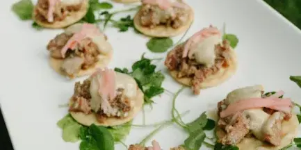 A white rectangular platter holding a row of gourmet appetizers topped with meat, cheese, and garnished with fresh greenery.