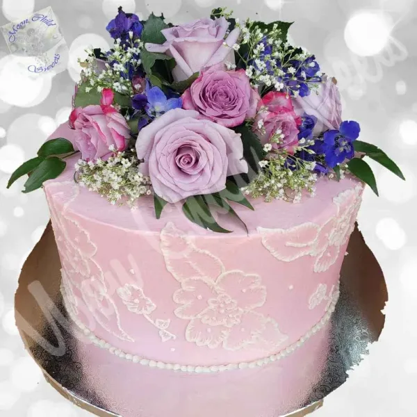 A pink frosted cake decorated with white lace patterns and topped with fresh pink roses and blue flowers.