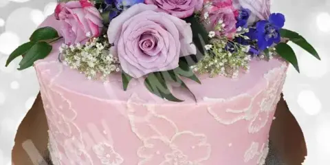 A pink frosted cake decorated with white lace patterns and topped with fresh pink roses and blue flowers.