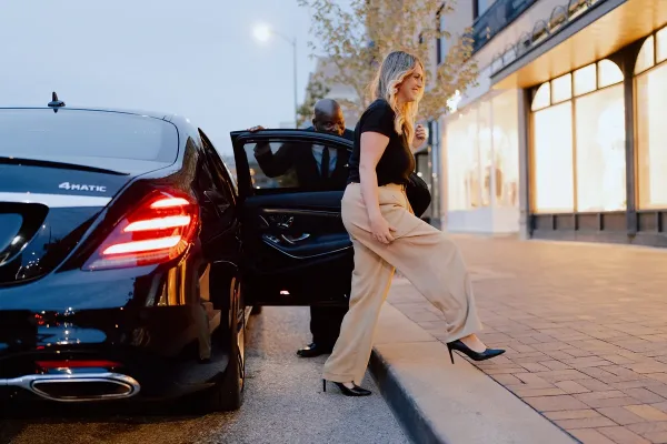 A professional chauffeur opens the rear door of a black luxury sedan for a female passenger to enter.