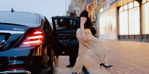 A professional chauffeur opens the rear door of a black luxury sedan for a female passenger to enter.