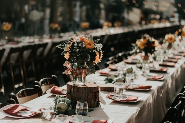 A long banquet table set with white linens, orange napkins, glassware, and rustic floral centerpieces in a wedding or event venue.