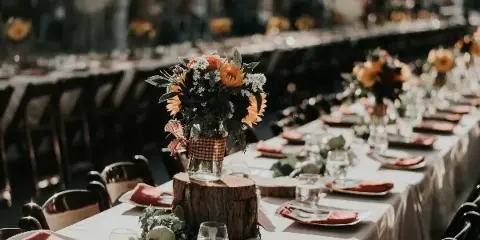 A long banquet table set with white linens, orange napkins, glassware, and rustic floral centerpieces in a wedding or event venue.
