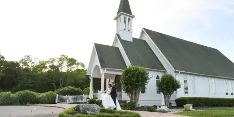 A bride and groom standing together in front of a white church with a steeple.