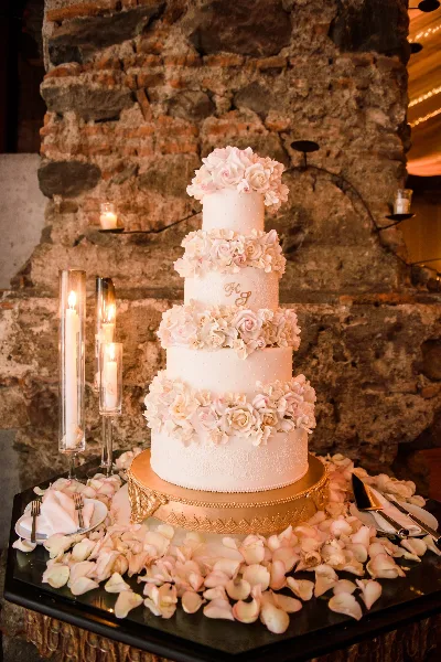A four-tier white wedding cake decorated with pink and white roses, displayed on a gold stand against a rustic stone wall.