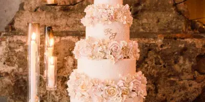 A four-tier white wedding cake decorated with pink and white roses, displayed on a gold stand against a rustic stone wall.