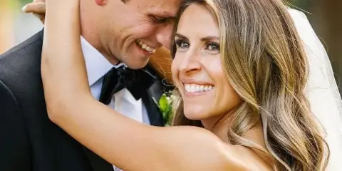 A smiling bride with styled hair and makeup embracing a groom in a tuxedo.