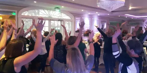 Guests dancing with raised hands at an indoor event with chandeliers and decorative lighting.