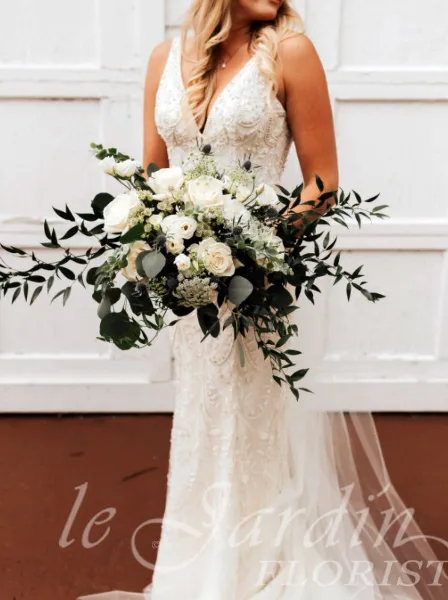A bride in a white lace gown holding a large bouquet of white roses and greenery.