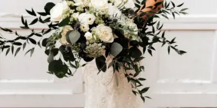A bride in a white lace gown holding a large bouquet of white roses and greenery.