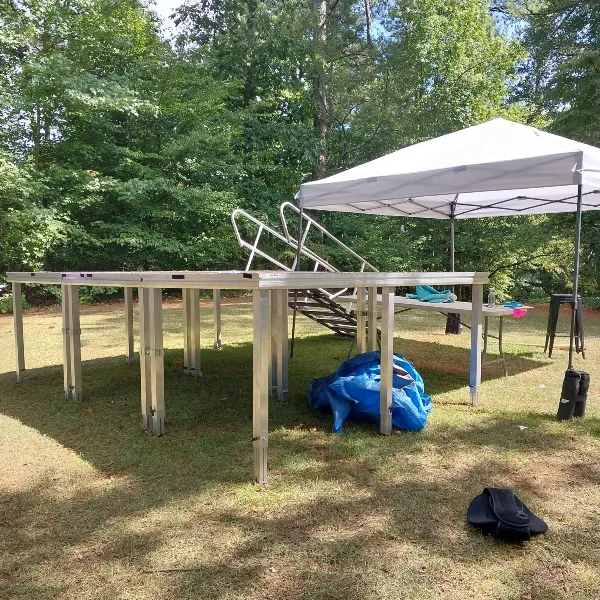 A modular aluminum stage platform with stairs set up on a grassy lawn next to a white canopy tent.