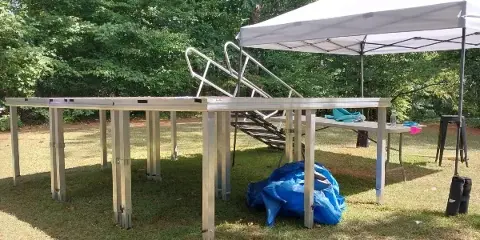 A modular aluminum stage platform with stairs set up on a grassy lawn next to a white canopy tent.
