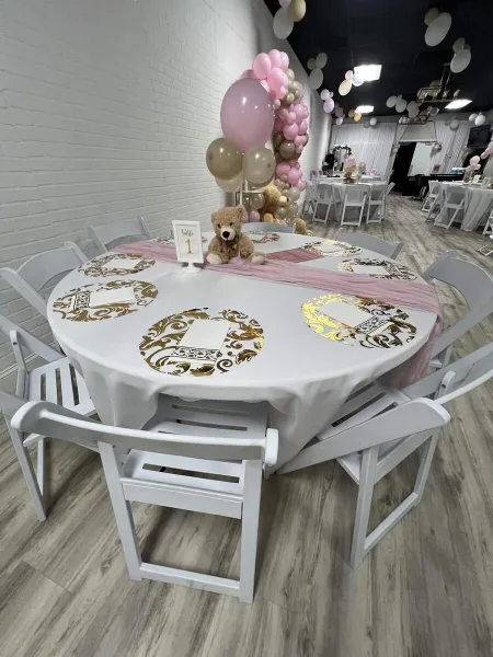 Interior of an event venue featuring a round table with white and gold tablecloth, white chairs, and a pink and gold balloon arch.