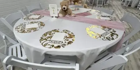 Interior of an event venue featuring a round table with white and gold tablecloth, white chairs, and a pink and gold balloon arch.
