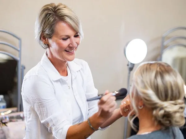 A makeup artist in a white shirt applying makeup to a client with an updo hairstyle in a studio.