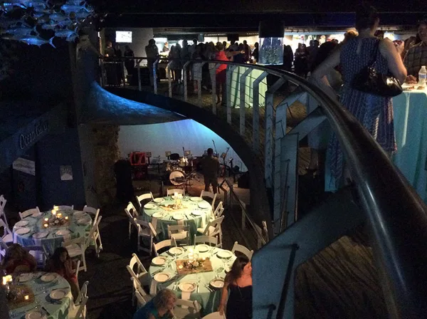 Indoor event reception with round tables set for a meal, guests seated, and a band on a small stage.