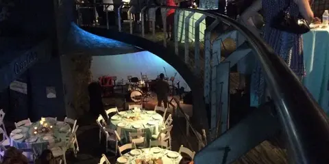 Indoor event reception with round tables set for a meal, guests seated, and a band on a small stage.