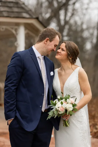 A bride and groom standing together outdoors, with the bride holding a bouquet of white and pink roses with eucalyptus.