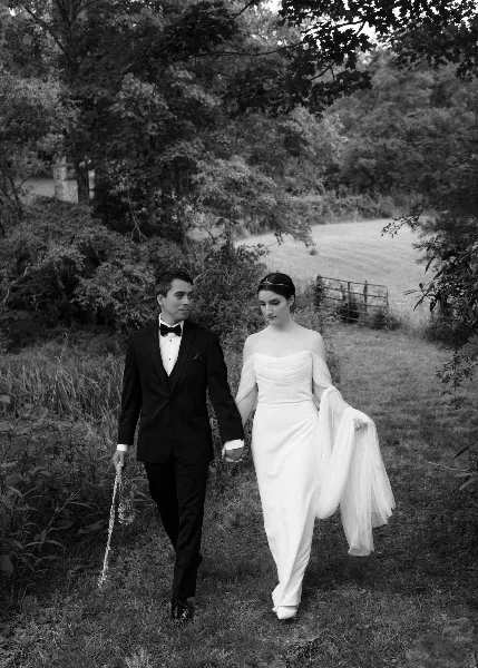 A black and white photograph of a bride and groom walking hand-in-hand through a grassy, wooded area.