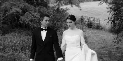 A black and white photograph of a bride and groom walking hand-in-hand through a grassy, wooded area.