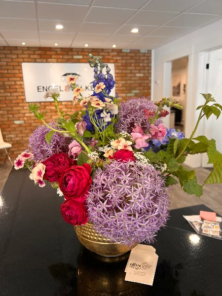 A vibrant floral arrangement with red ranunculus, purple allium, and delphinium in a gold bowl, displayed on a studio table with an Olive MaGoo's business card.