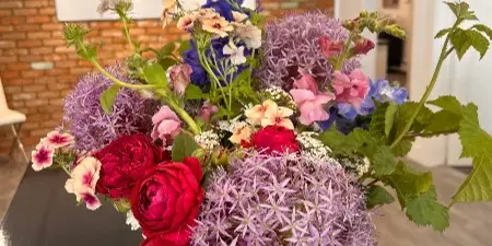 A vibrant floral arrangement with red ranunculus, purple allium, and delphinium in a gold bowl, displayed on a studio table with an Olive MaGoo's business card.