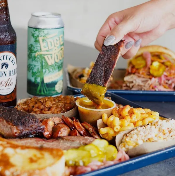 A barbecue platter with smoked meats, beans, mac and cheese, and a pulled pork sandwich, with a hand dipping a slice of meat in sauce.