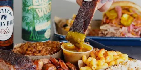 A barbecue platter with smoked meats, beans, mac and cheese, and a pulled pork sandwich, with a hand dipping a slice of meat in sauce.