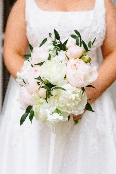 A bride in a white lace dress holding a bouquet of white and pale pink peonies with greenery.