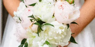 A bride in a white lace dress holding a bouquet of white and pale pink peonies with greenery.