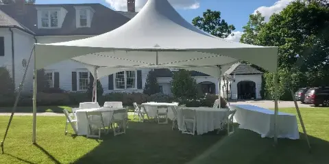 A white event tent set up on a grassy lawn with white round tables and chairs arranged underneath.